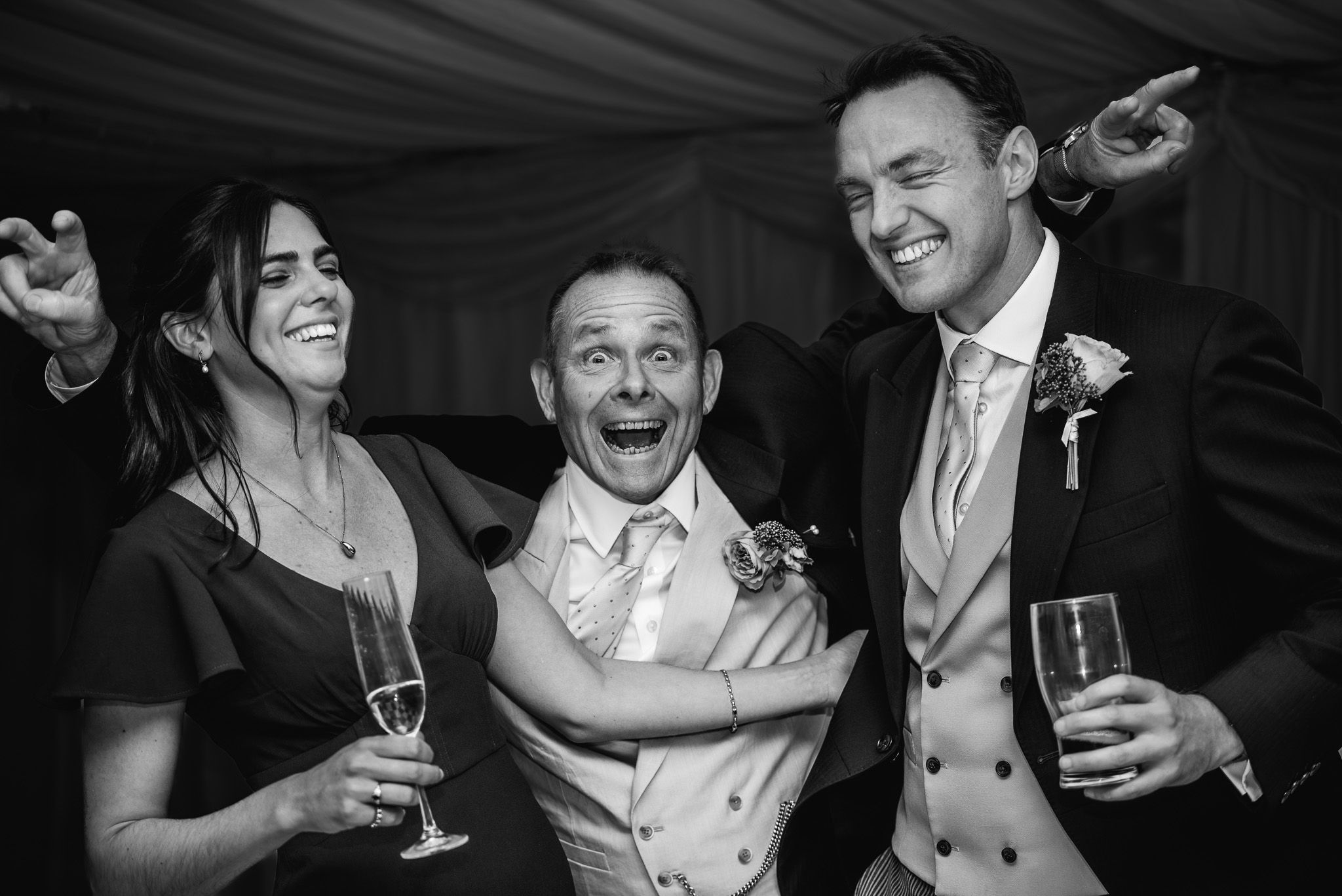 Wedding party laughing and celebrating with champagne glasses in formal attire at reception.