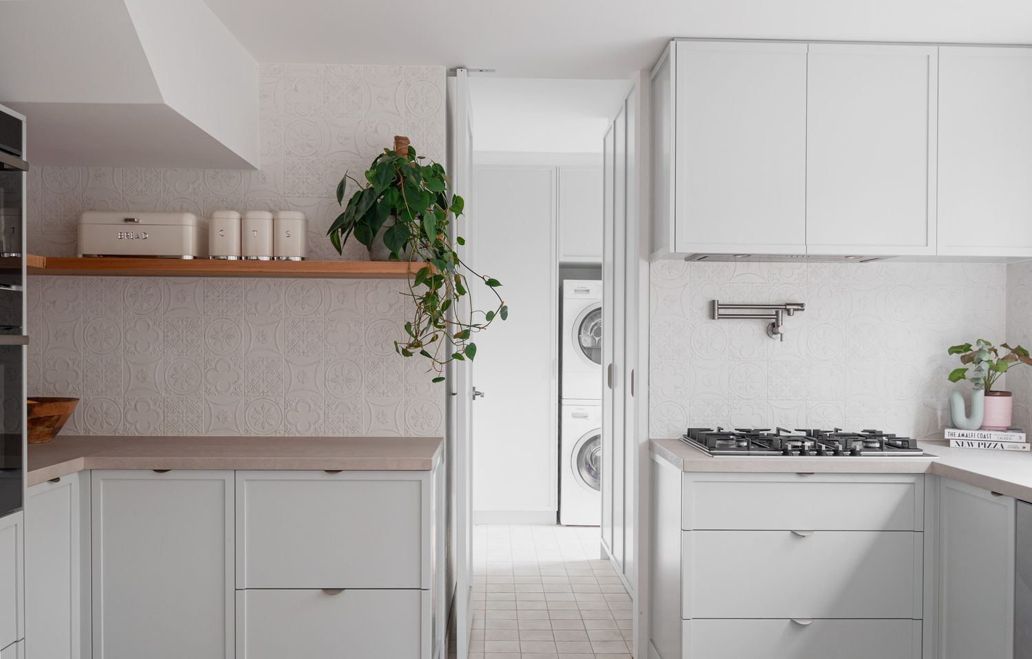 White modern kitchen with wooden shelving, integrated appliances, and trailing indoor plants.