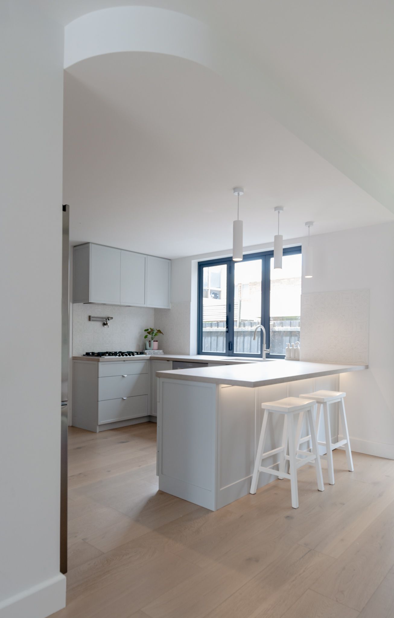 Modern white kitchen with light wood floors, blue-framed windows, and a minimalist island with bar stools.