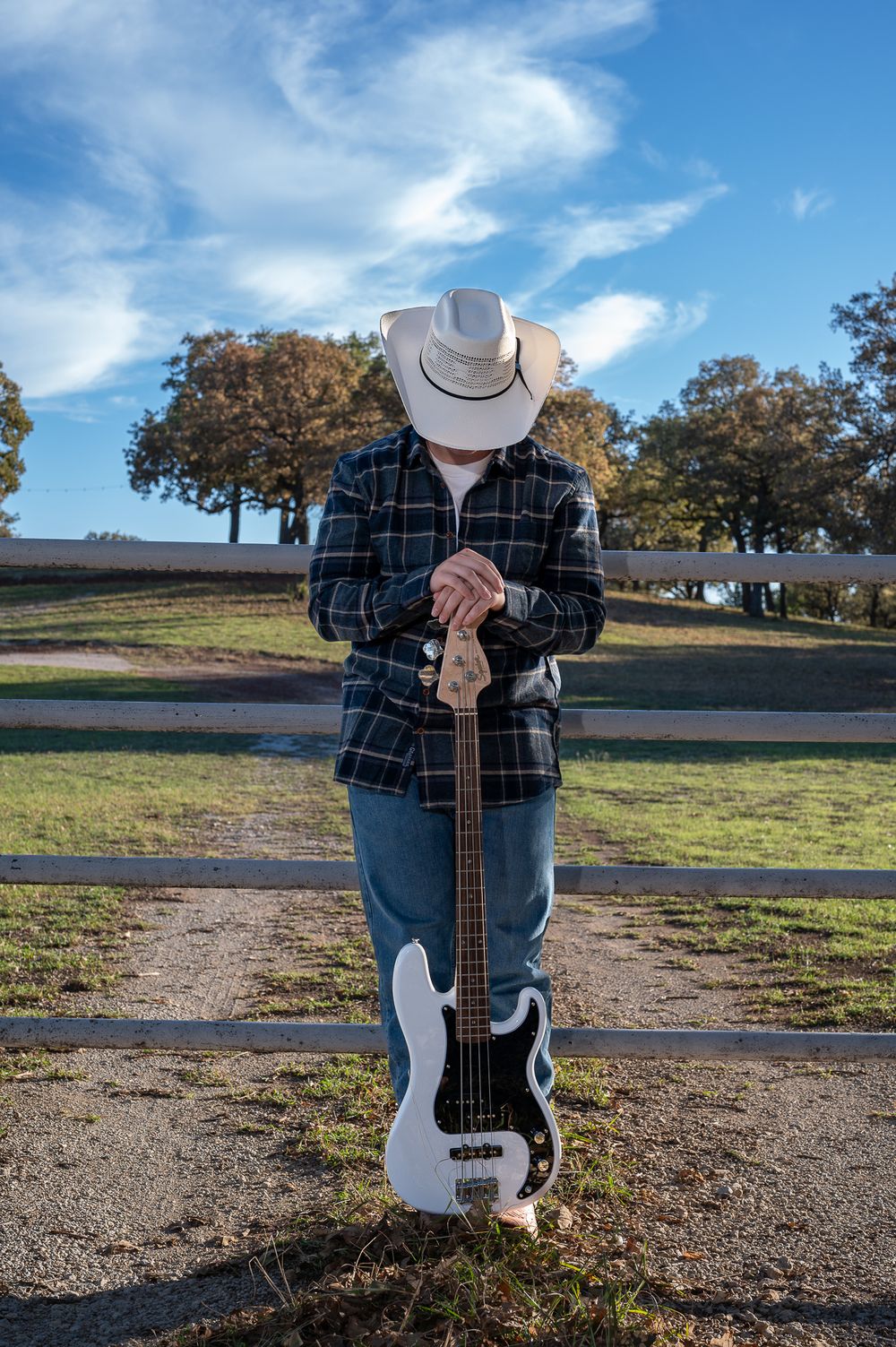 A teenage musician in plaid shirt and cowboy hat poses with bass guitar on dirt path.