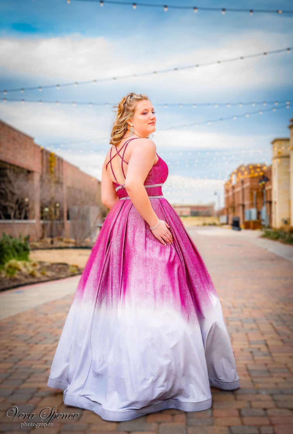 A teenager in an ombre pink to white formal gown poses on brick path under string lights.
