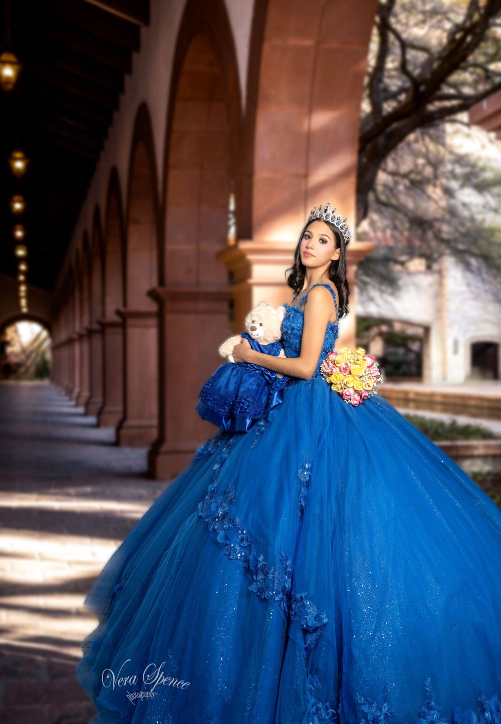 Quinceañera portrait in sparkling blue ball gown with crown under archway.