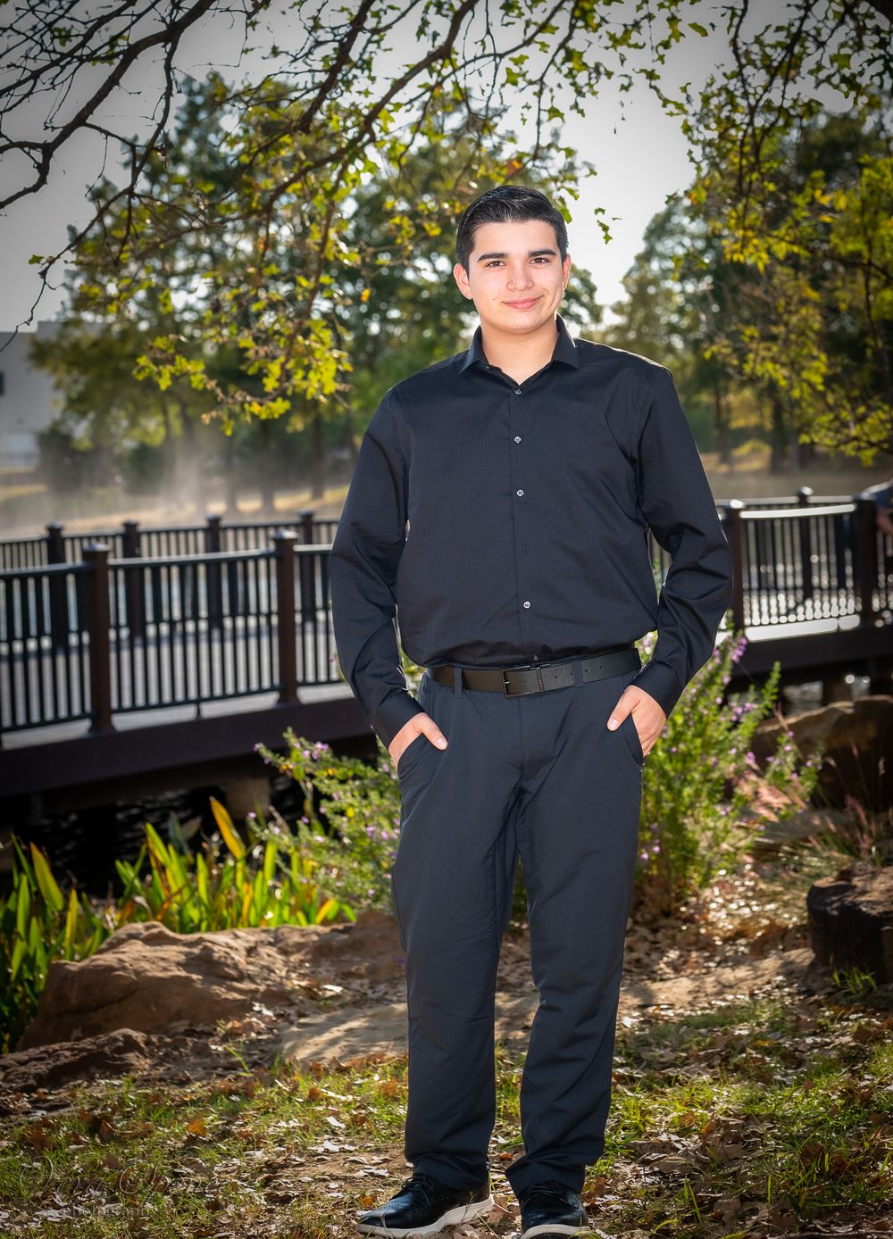 A person in business casual attire stands in front of a decorative railing and trees.