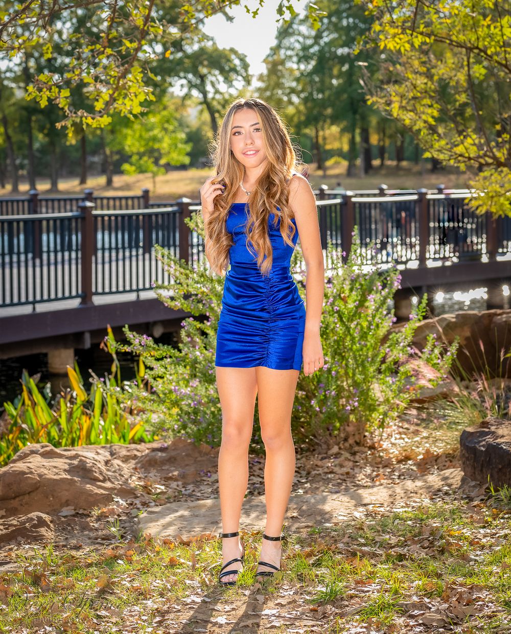 Teen in blue dress poses outdoors by a wooden bridge on a sunny day.