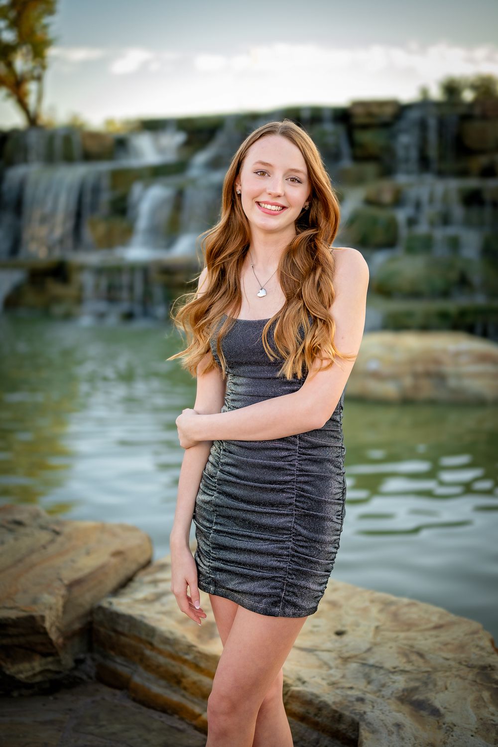 A teenager in ruched gray dress poses by cascading waterfalls.