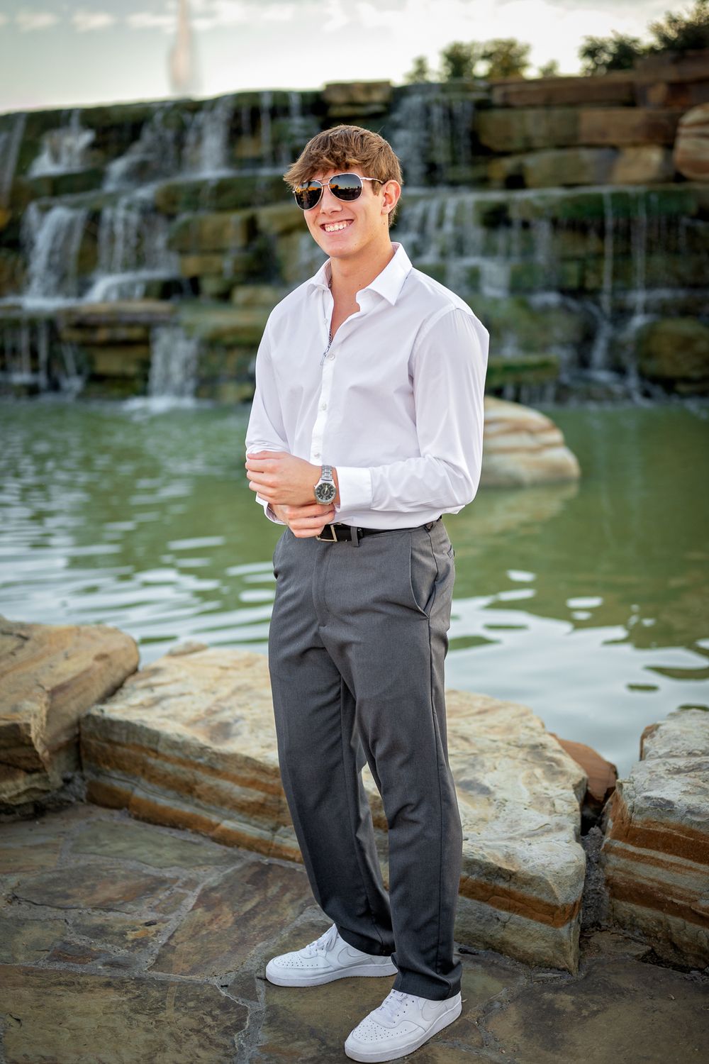 A teenager in white dress shirt and gray pants smiles by tiered waterfalls.
