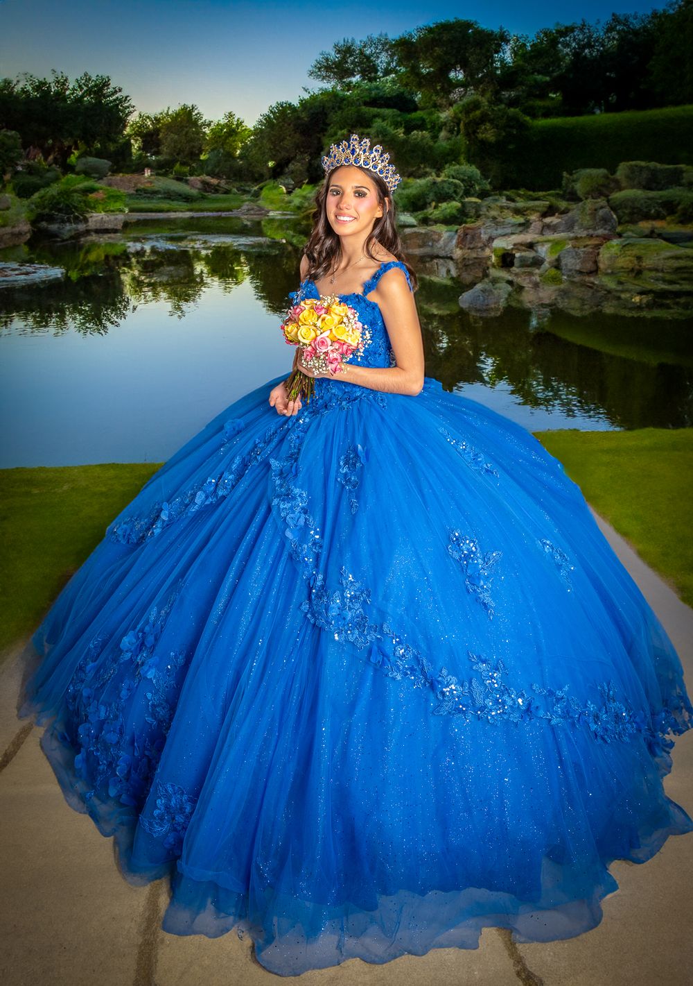 A teenager in royal blue quinceañera ball gown poses by scenic pond at sunset.