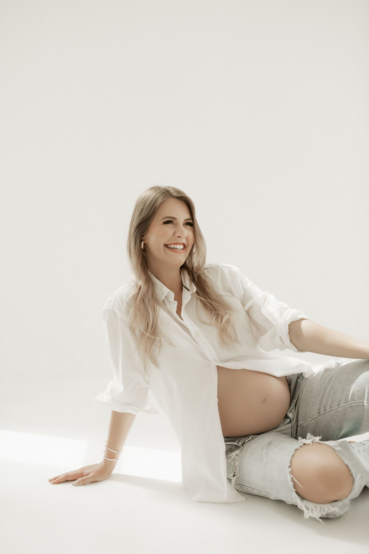 Casual maternity portrait in white shirt and distressed jeans on white background.
