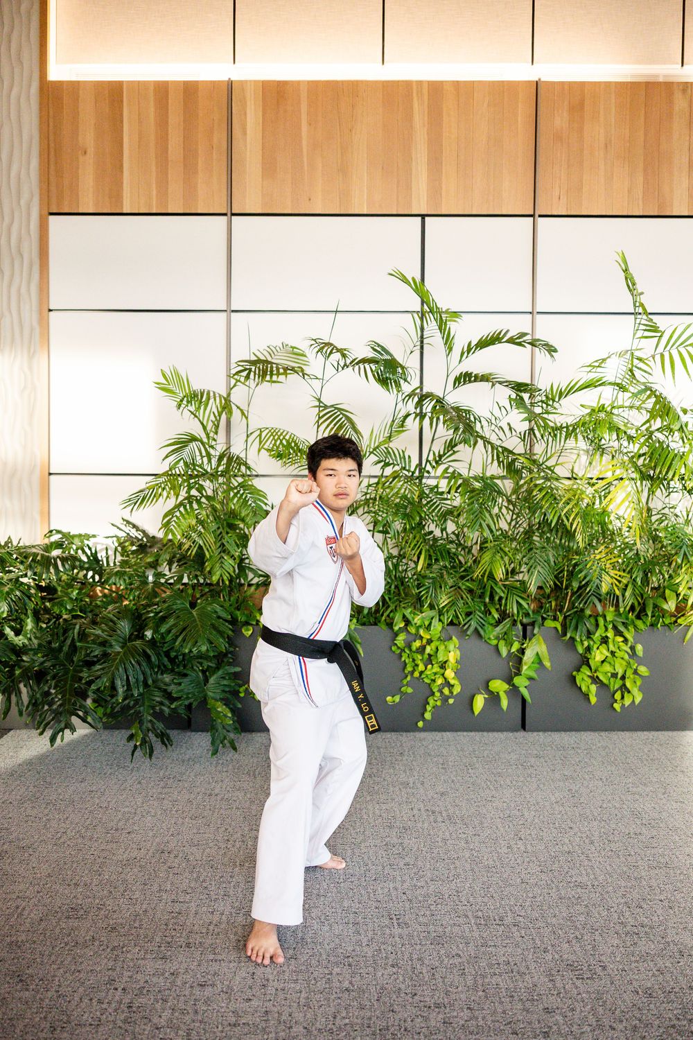 Martial artist in white uniform performs dynamic kicks and poses in sequence against gray wall with green plants.