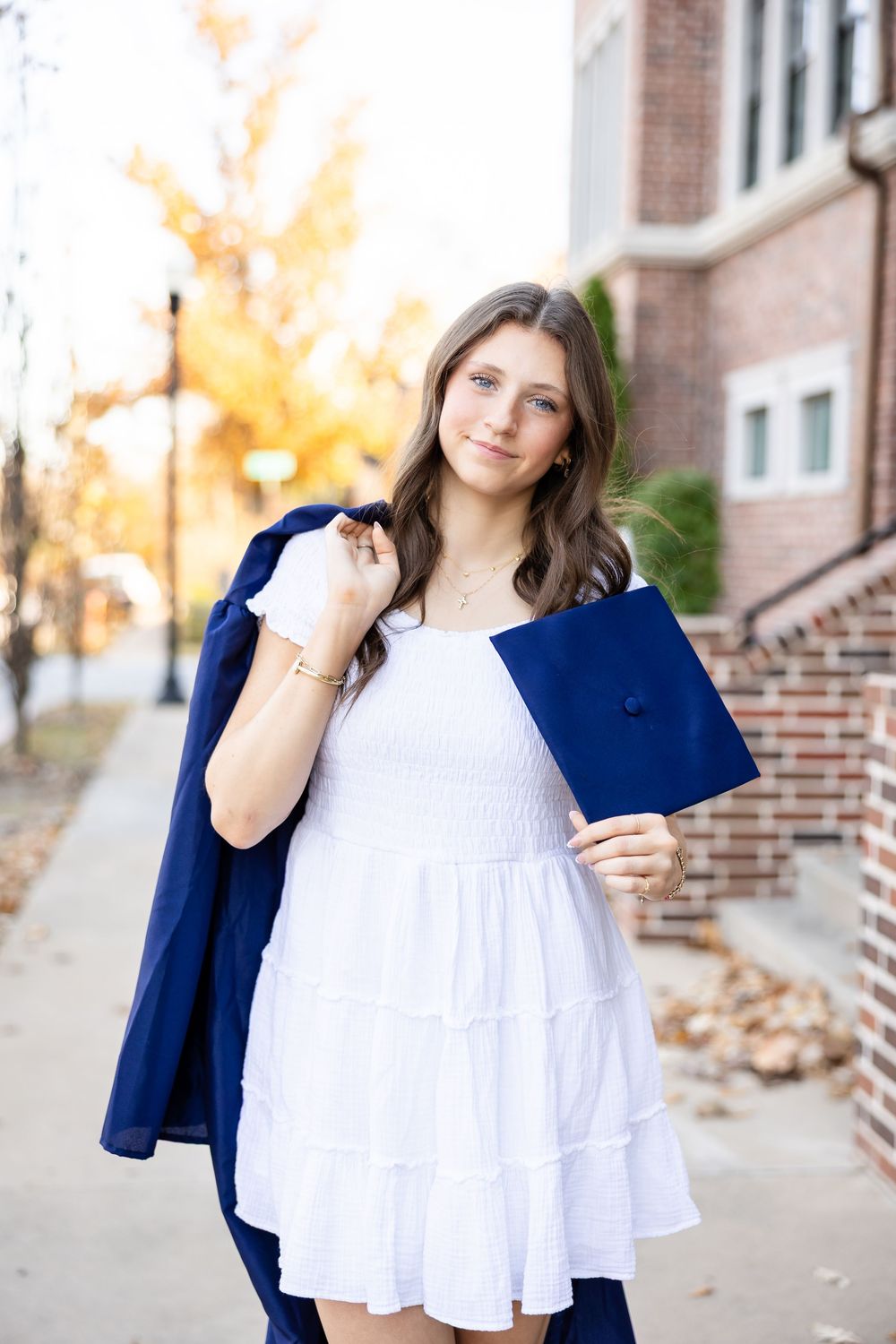 Graduate in white dress and navy blue cap and gown stands outside brick building on autumn day.