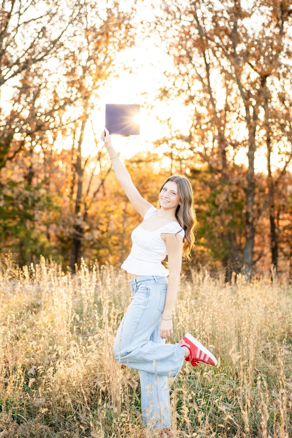 A person in jeans and white top holds up a diploma while standing in a golden autumn field at sunset.