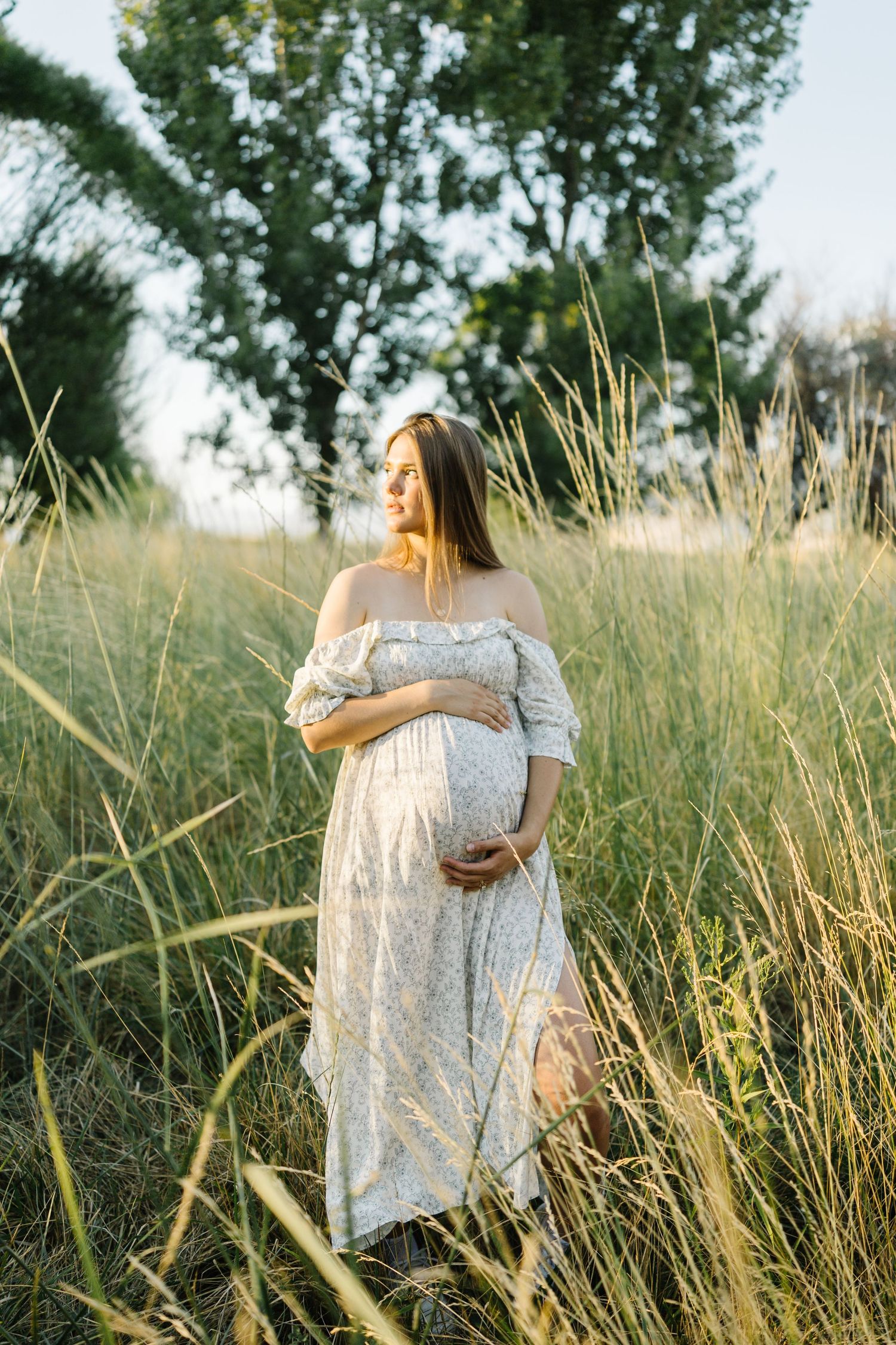Ethereal maternity photo in a sunlit field of tall grass featuring an off-shoulder white lace dress at golden hour.