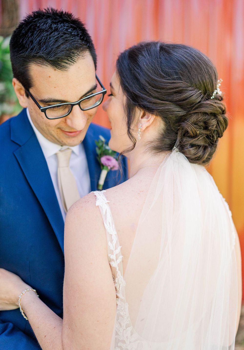 Intimate wedding moment captured with navy suit and bridal veil against coral backdrop.