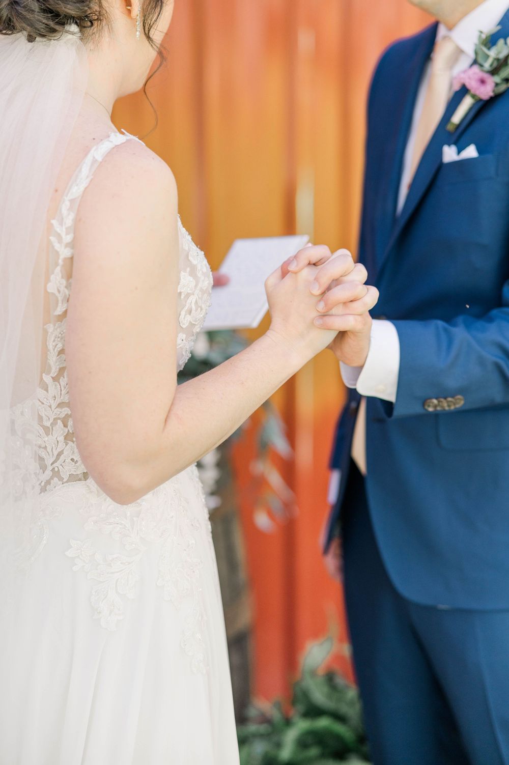 Close-up of couple holding hands during wedding ceremony against orange wall.