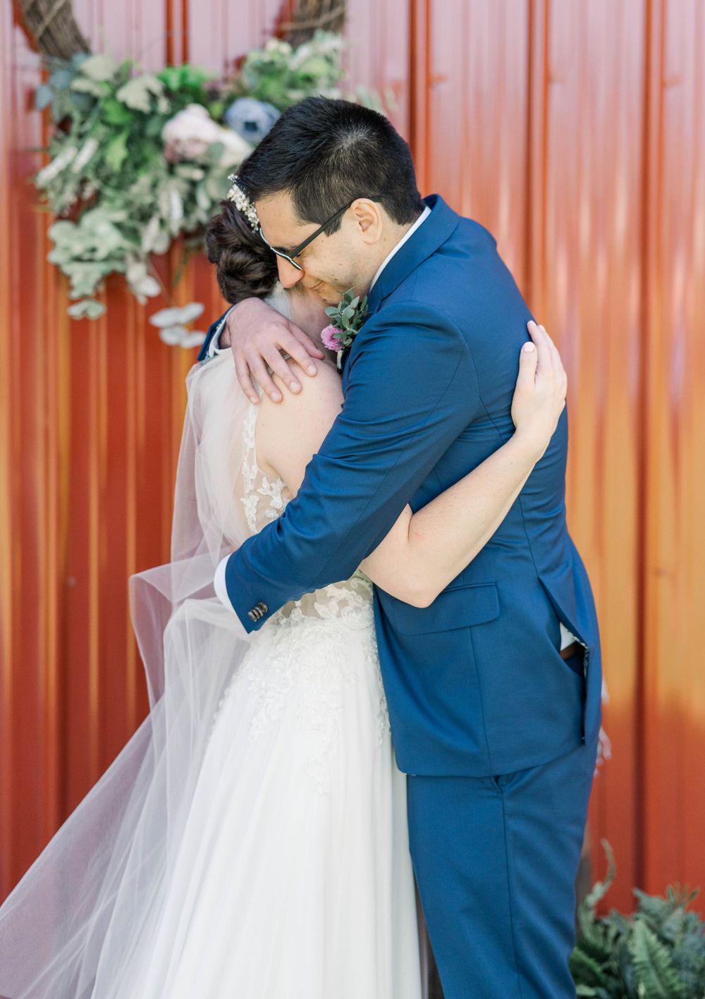 A couple embraces tenderly against a coral-colored wall decorated with white floral arrangements.