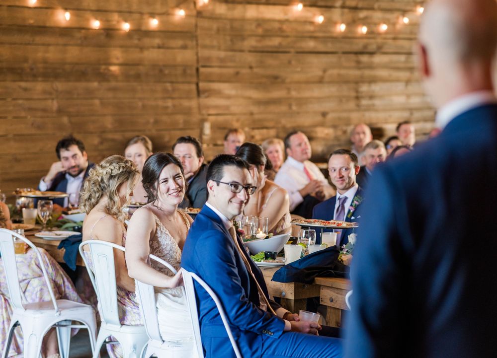 Wedding reception guests seated at long wooden tables under string lights in rustic venue.