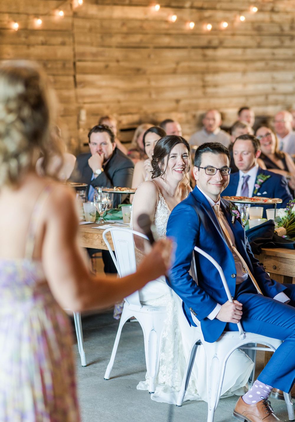 Wedding guests listen intently to a speech during an intimate barn reception with string lights.