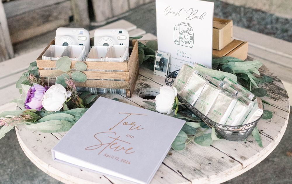 Wedding guest book and favor display with white boxes and greenery on rustic wooden table.