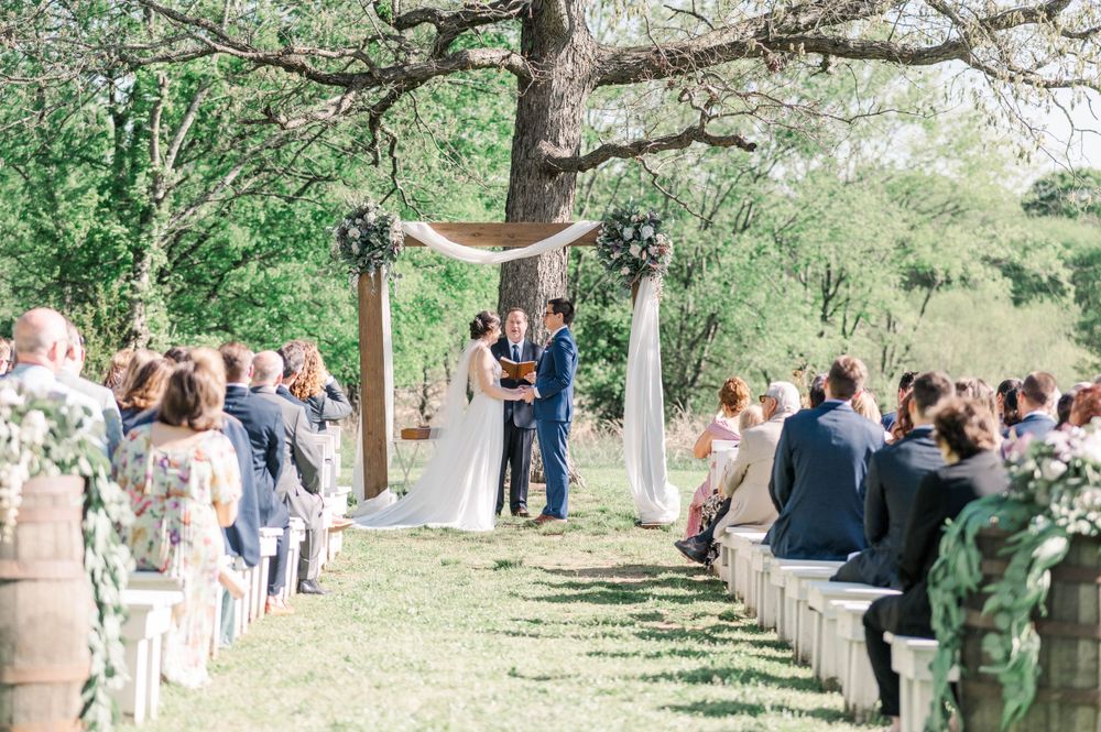 Rustic outdoor wedding ceremony with wooden arch and white draped fabric among trees.