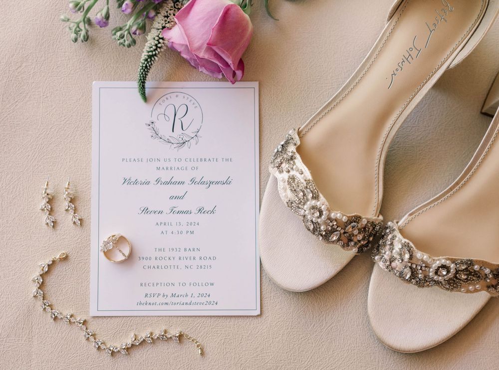 Wedding invitation with monogram design displayed with bridal shoes and jewelry. The 1932 Barn Venue, Charlotte, NC