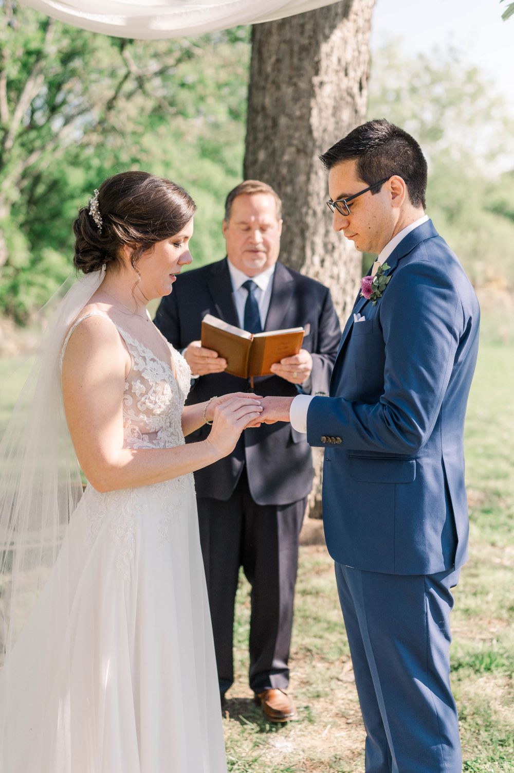 Wedding ceremony with couple exchanging rings during vows under a tree.