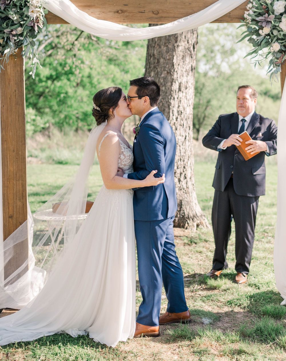 Wedding ceremony under white draped arch with couple sharing first kiss.