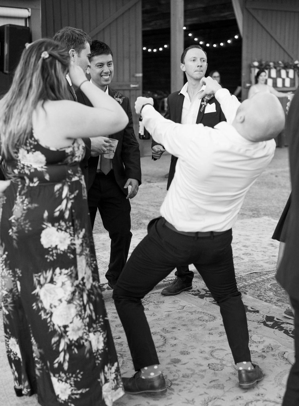 Wedding guests dance energetically at a barn reception in black and white photography.