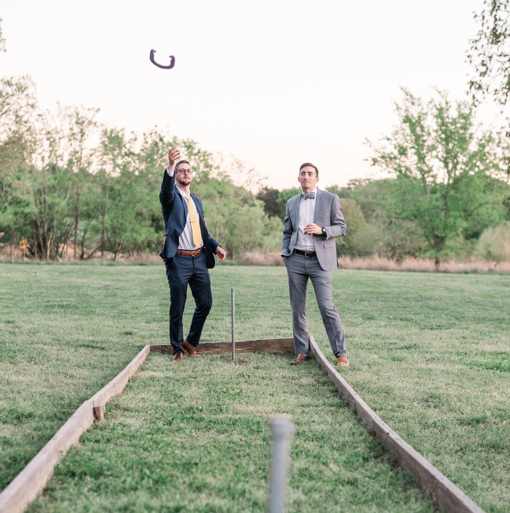 Two men in suits play horseshoes on a grassy field near railroad tracks.