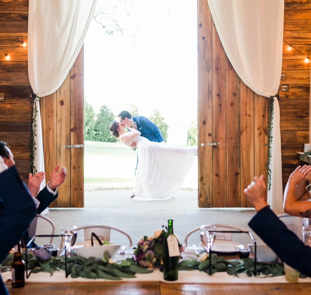 Newlyweds share a romantic kiss in a rustic barn doorway framed by white curtains.
