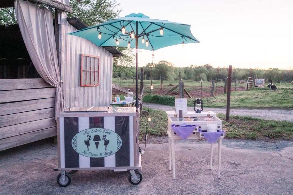 Rustic outdoor event setup with a turquoise umbrella and vintage cart against a sunset backdrop.