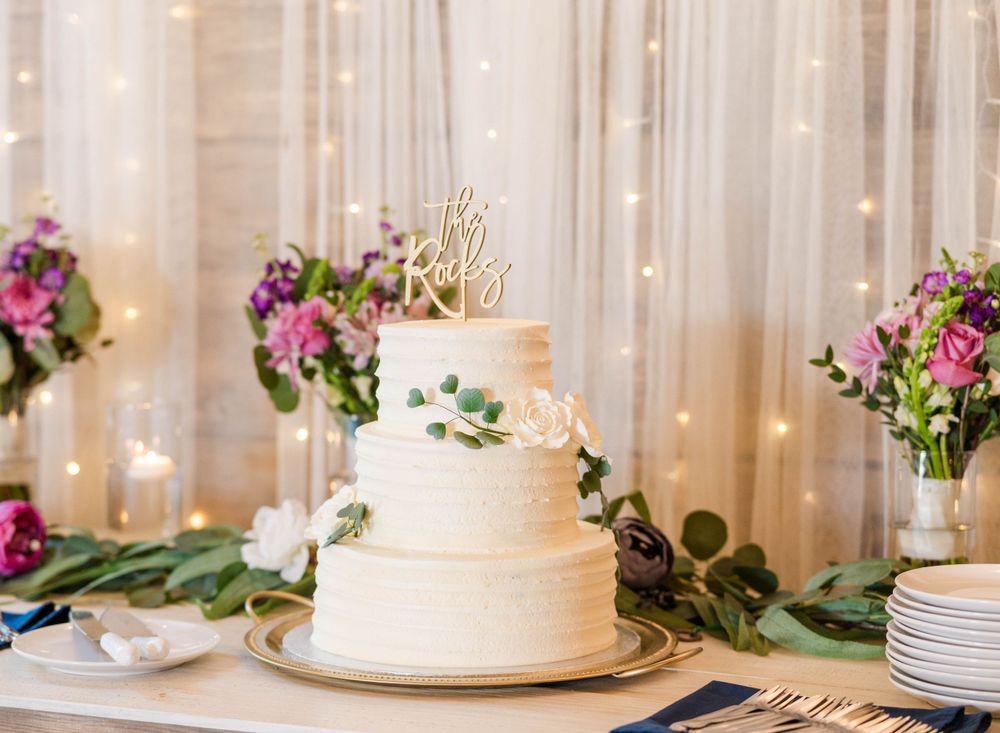 Three-tiered white wedding cake decorated with greenery and flowers sits beside floral centerpieces.