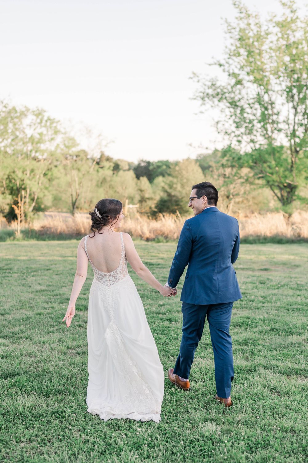 A newlywed couple walks hand in hand across a sunlit grassy field in elegant wedding attire.