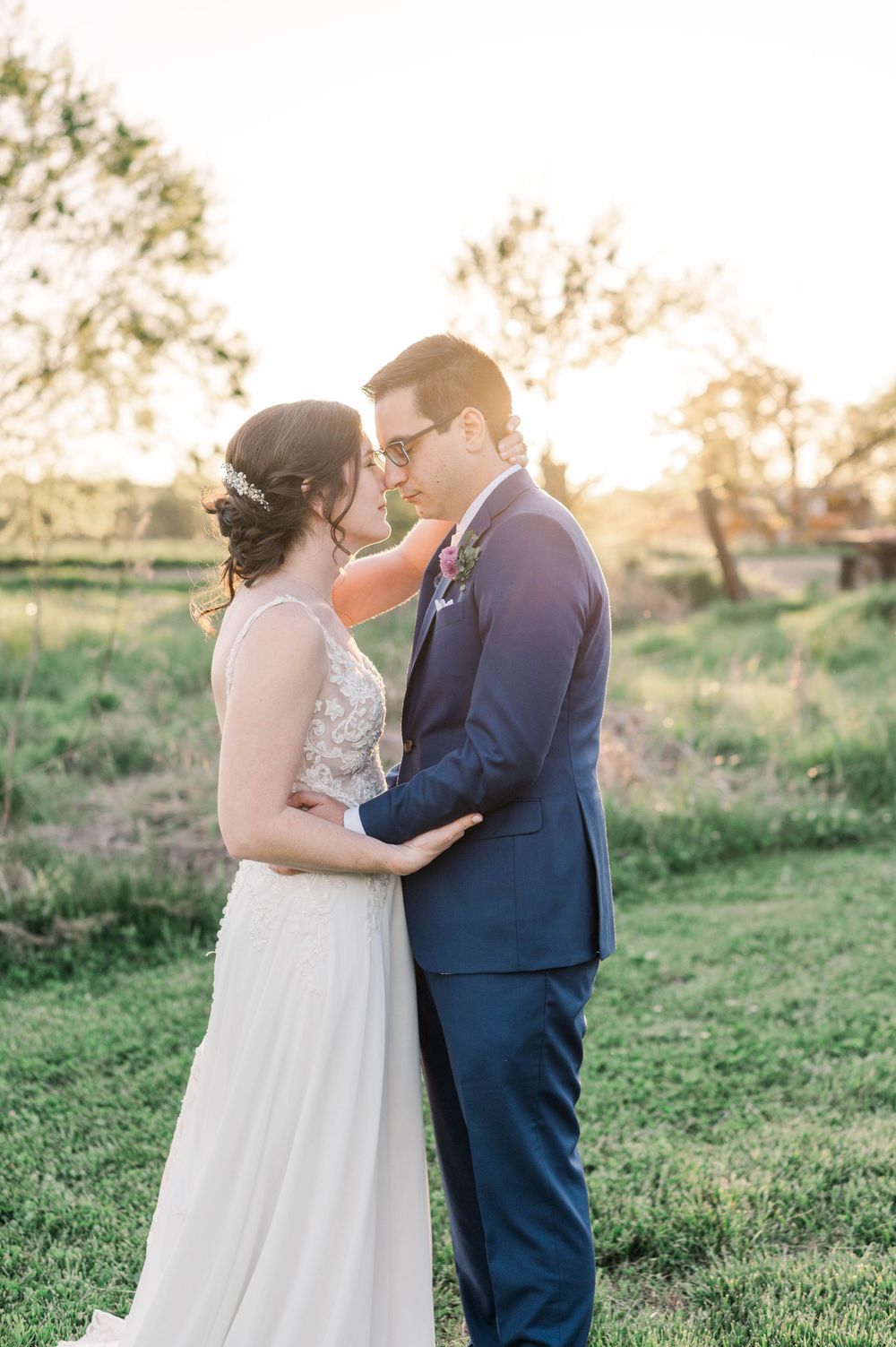 A romantic wedding couple embraces in a golden-hour meadow wearing navy suit and lace gown. The 1932 Barn Venue, Charlotte, NC