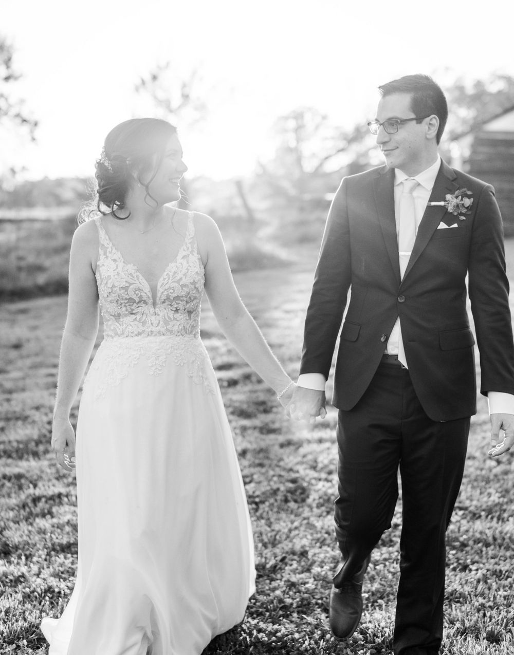Black and white portrait of newlyweds walking and smiling in an outdoor setting. The 1932 Barn Venue, Charlotte, NC