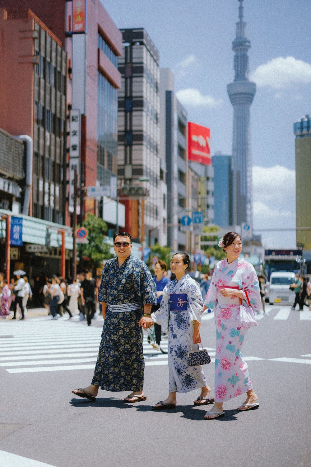 Three people wearing traditional yukata kimonos crossing street with Tokyo Skytree visible.