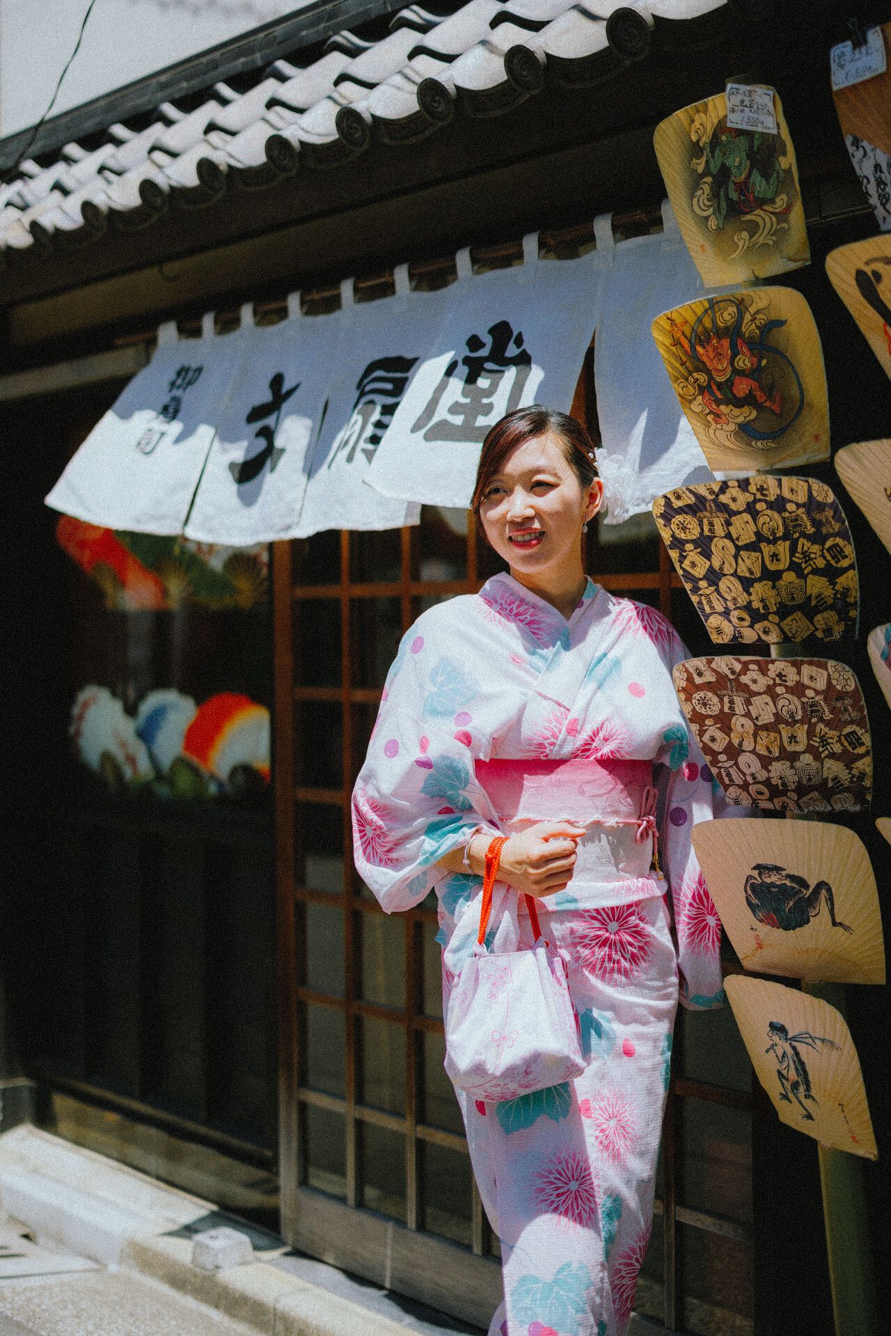 Person in pink floral yukata poses outside traditional Japanese storefront with hanging banners.