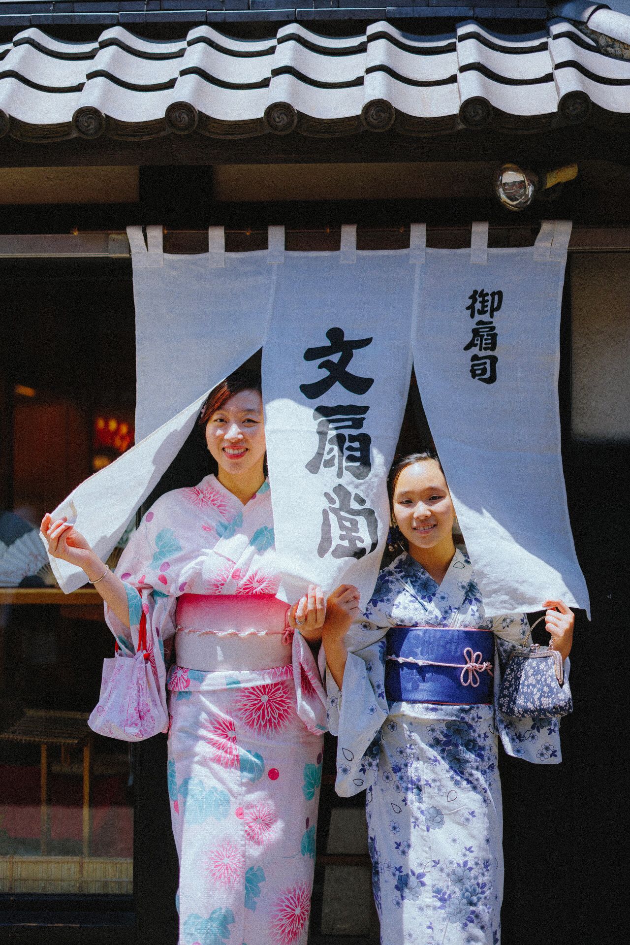 Two people in colorful yukatas pose beneath traditional Japanese shop curtains and signage.