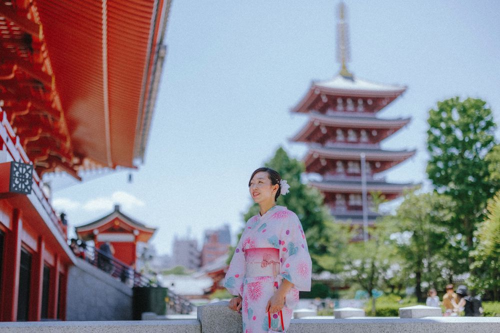 Person in pink floral yukata standing in front of red Japanese pagoda on sunny day.