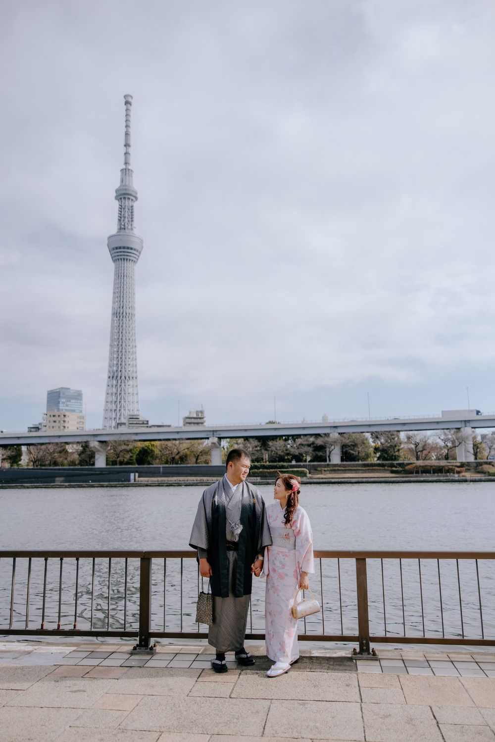 Two people sit on a bench overlooking a river with the Tokyo Skytree tower visible in the background.