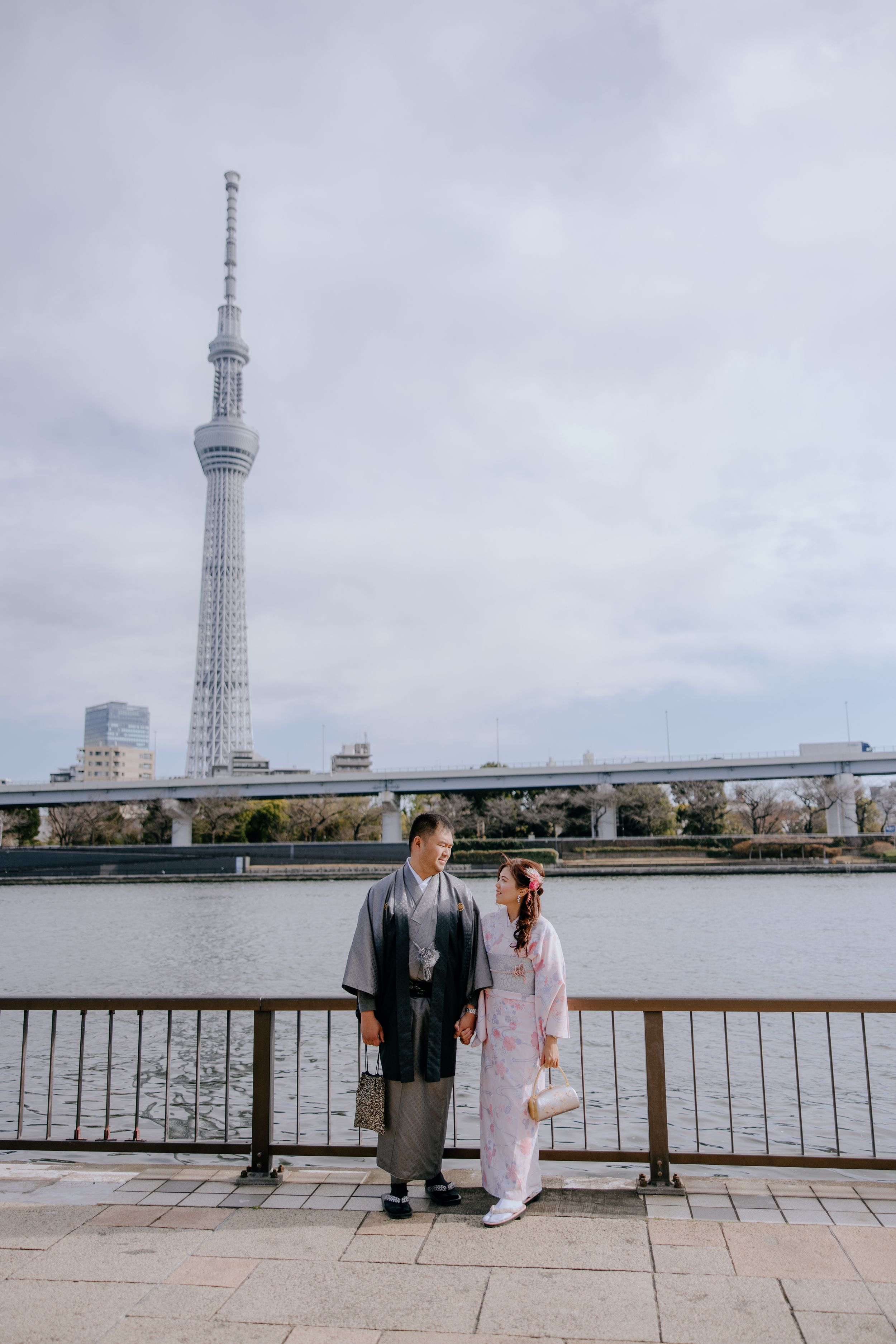 Two people sit on a bench overlooking a river with the Tokyo Skytree tower visible in the background.