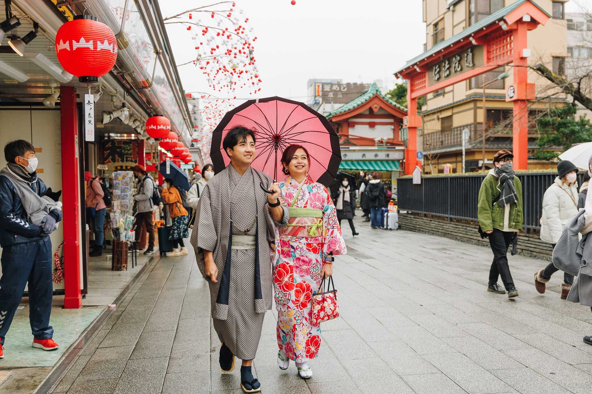Couple in traditional Japanese attire walking under red lanterns with a pink umbrella.