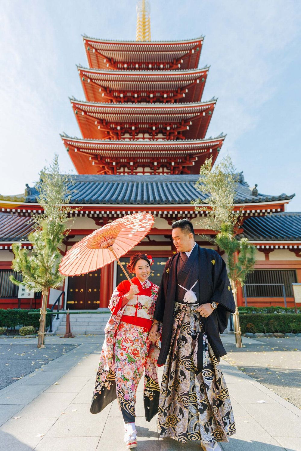 Couple in traditional Japanese dress standing before a red pagoda with parasol.