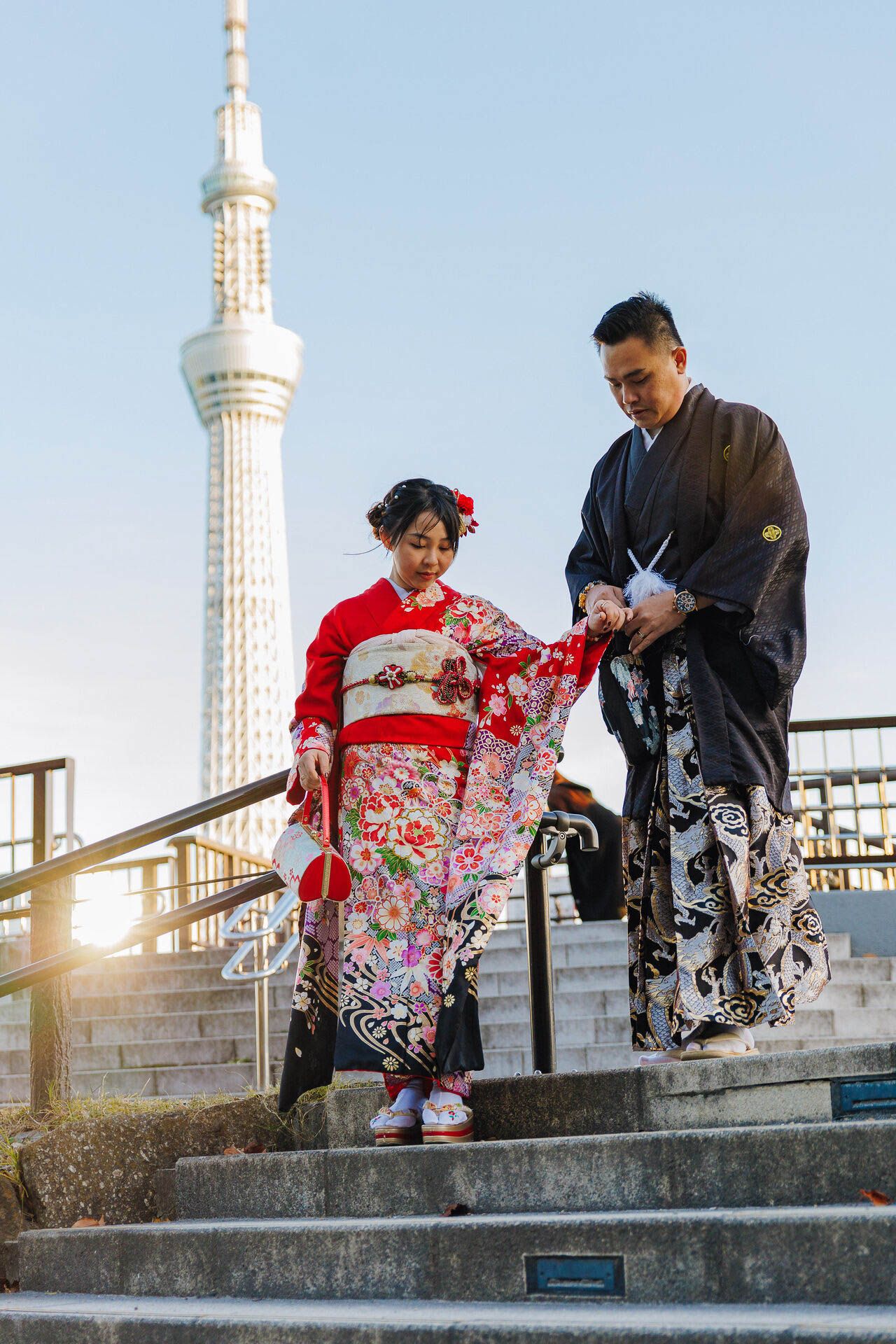 A couple in traditional Japanese kimonos walking down steps with Tokyo Skytree tower in background at sunset.