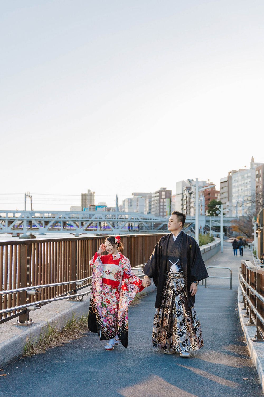 A couple in traditional kimonos walking along a riverside path with city buildings and bridge in background.