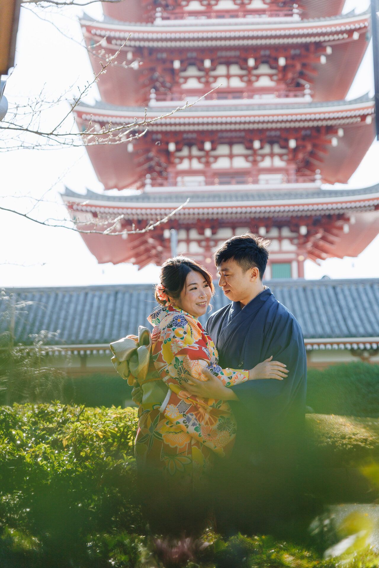 Couple in traditional Japanese dress embracing near red pagoda temple in spring garden setting.