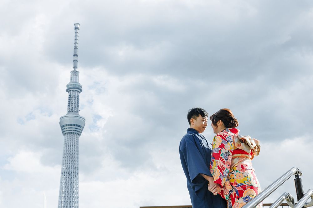 A couple in traditional Japanese attire pose together with Tokyo Skytree tower against cloudy sky.