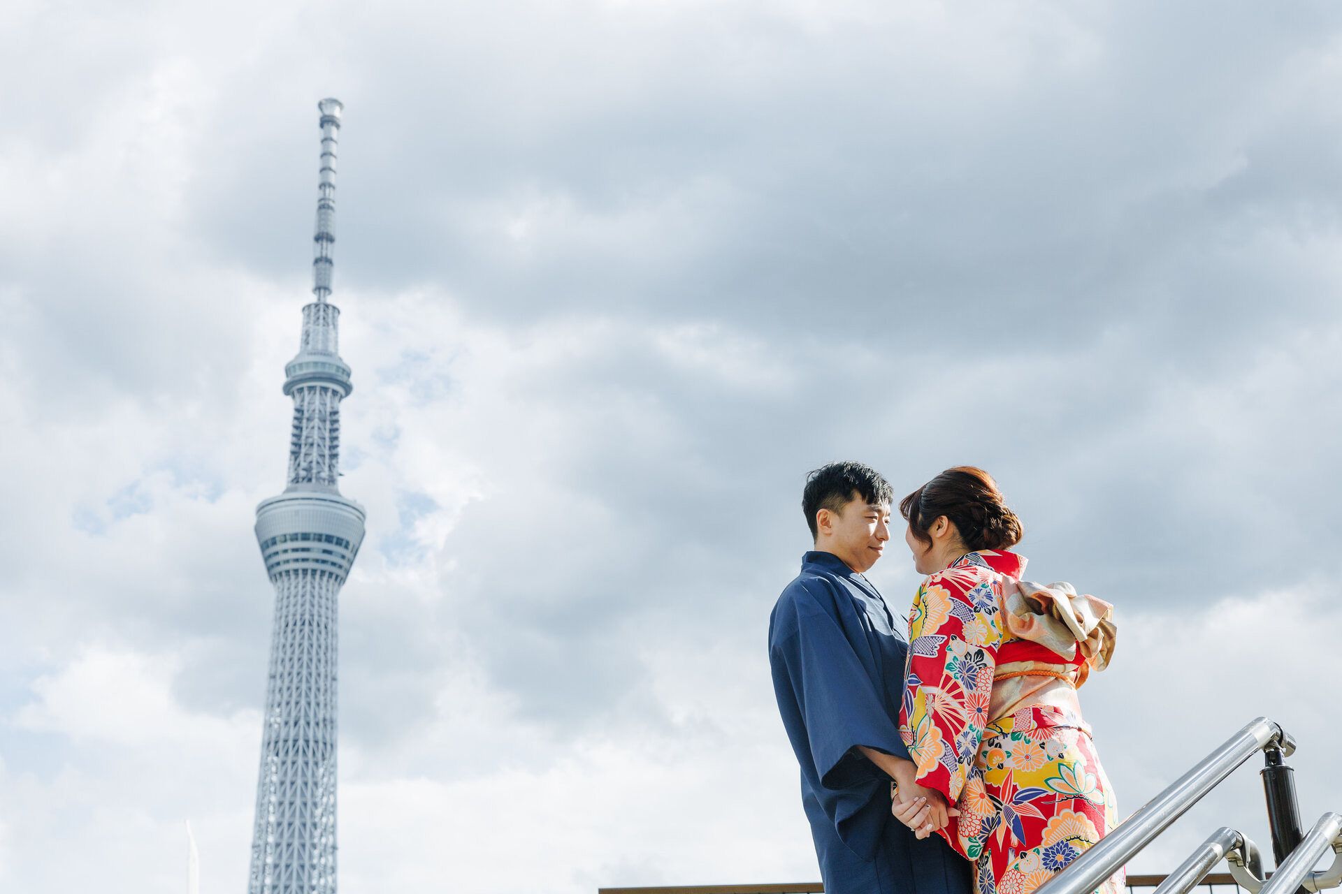 A couple in traditional Japanese attire pose together with Tokyo Skytree tower against cloudy sky.