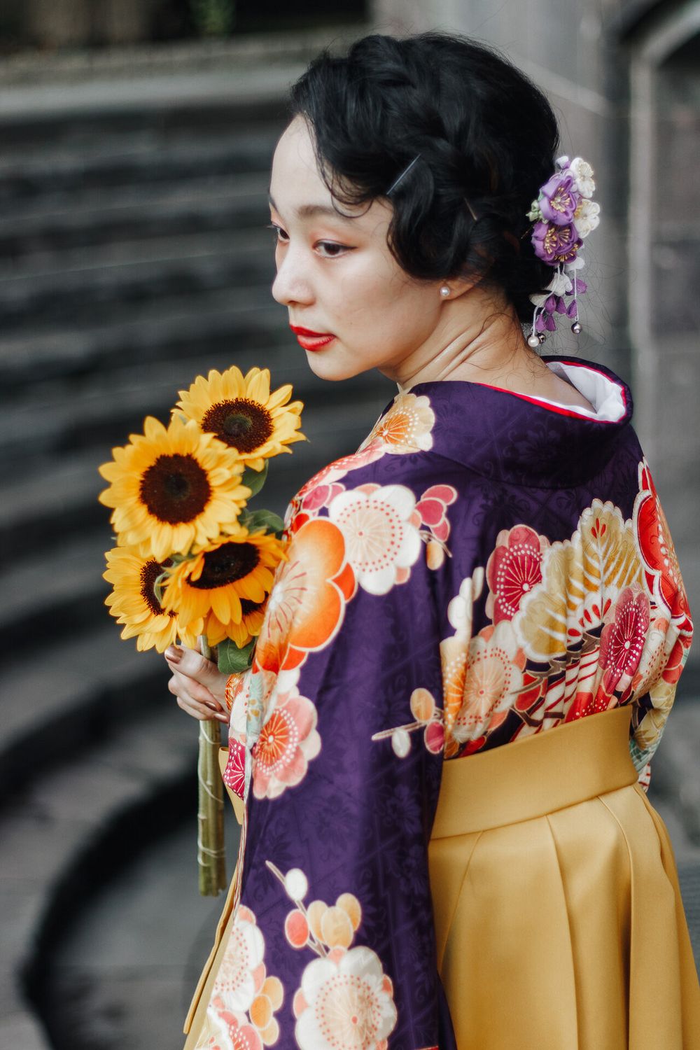 Traditional kimono with purple floral pattern and sunflowers against stone wall backdrop.