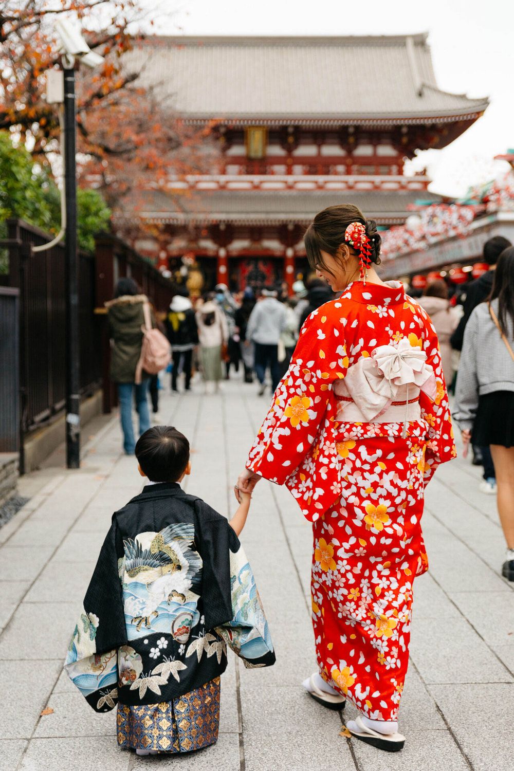 Traditional Japanese scene with people wearing kimonos walking along a temple pathway lined with autumn trees.