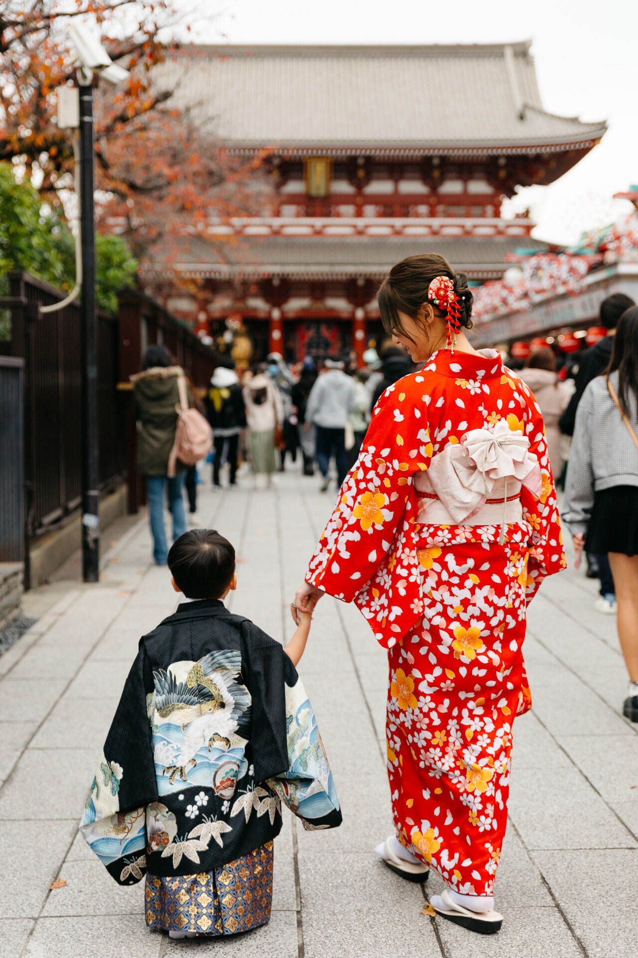Traditional Japanese scene with people wearing kimonos walking along a temple pathway lined with autumn trees.